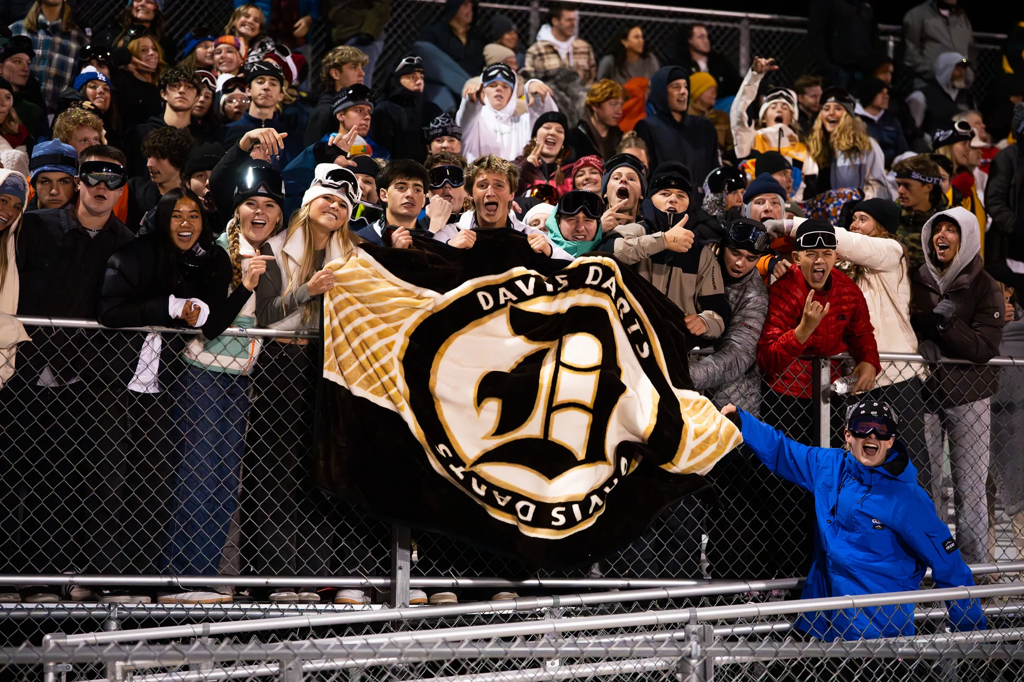 A packed high school student section on a cold night holding up a black-and-gold custom mascot blanket over the fence